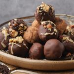 A plate with an assortment of vegan hazelnut truffles, some decorated with nuts and white glaze, others dusted with cocoa powder.
