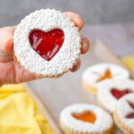 A hand holds a vegan linzer cookie with a powdered sugar coating and heart-shaped red filling. More cookies are in the background.
