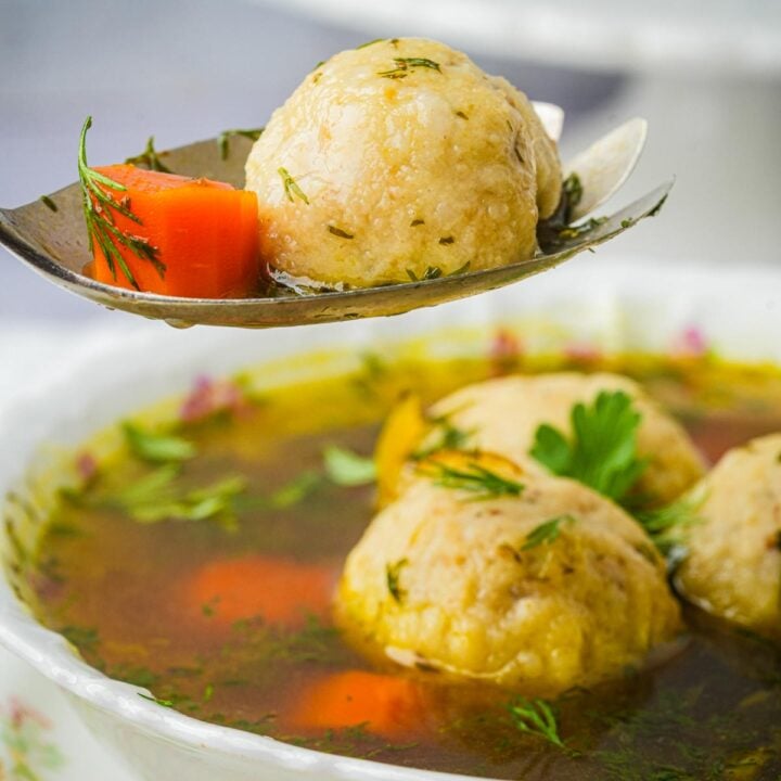 A spoon holding a vegan matzo ball and carrot above a bowl of soup with matzo balls, garnished with herbs.