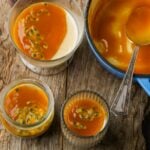 Three panna cotta desserts topped with passion fruit sauce on a wooden surface, with an additional serving of sauce in a blue dish with a spoon.