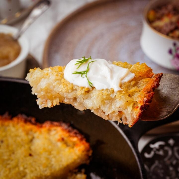 A slice of vegan potato kugel on a spatula, topped with a dollop of sour cream and a sprig of dill. Other food items are blurred in the background.