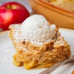 Slice of vegan apple kugel topped with a scoop of vanilla ice cream on a plate, with a red apple and a baking dish in the background.