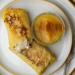 Two cooked vegan blintzes on a plate, one partially eaten, next to a small glass bowl of applesauce topped with cinnamon. A fork is on the side.