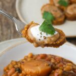 A fork holding a slice of vegan kishke with gravy, vegan sour cream and garnish over a bowl of vegan cholent.