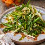 A plate of Korean scallion salad with sliced green onion, red pepper flakes, and sesame seeds. A bulb of garlic is nearby.