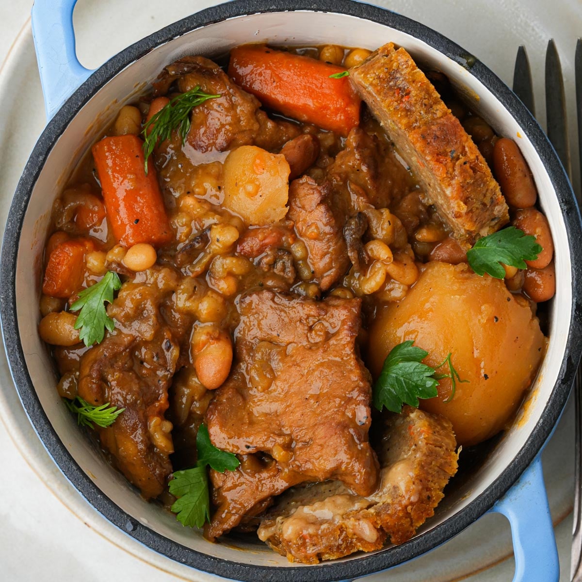 Vegan cholent in a round blue pot containing seitan, carrots, potatoes, beans, and garnished with parsley.