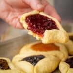 A hand holds a triangular vegan hamantaschen filled with red jam above a tray of assorted pastries with various fillings.