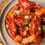 A plate of vegan Korean fried chicken coated in red sauce, garnished with sesame seeds, chopped green onions, and accompanied by cubed vegetables.
