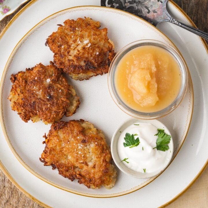 Three fried vegan latkes on a plate with a small bowl of applesauce and a dollop of sour cream garnished with parsley.