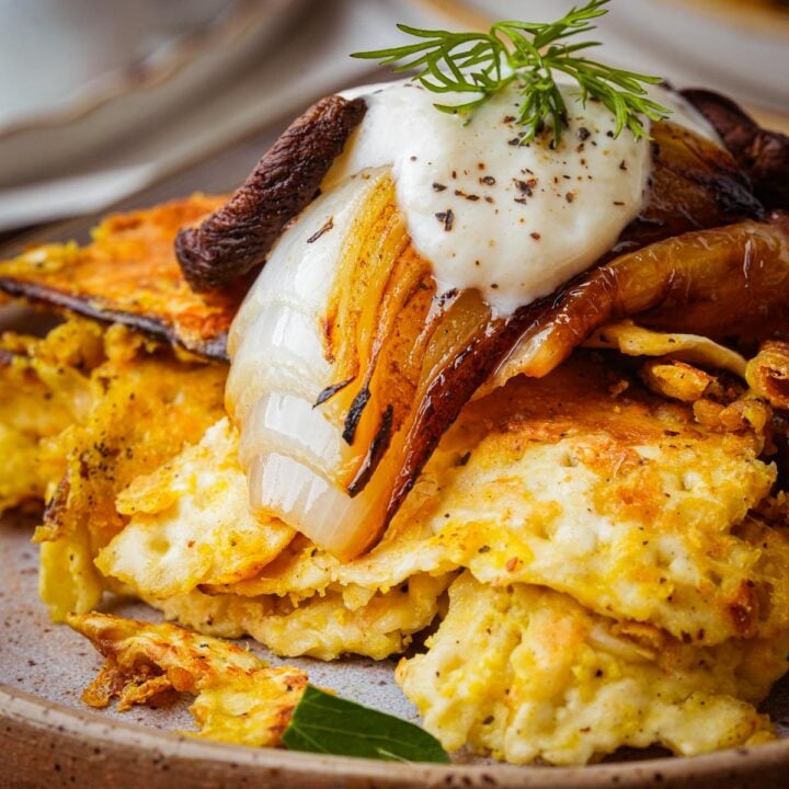 A stack of crispy vegan matzo brei topped with caramelized onions, a dollop of sour cream, fresh dill, and ground pepper on a brown plate.