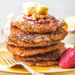 A stack of vegan matzo meal pancakes topped with butter, syrup, and powdered sugar, accompanied by a strawberry on a plate with a fork.
