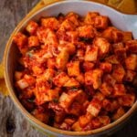 A bowl of vegan radish kimchi, seasoned with red pepper paste sits on a wooden table.