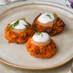 Three vegan sweet potato latkes topped with applesauce and cream on a beige plate, garnished with dill sprigs.