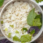 Bowl of coconut jasmine rice garnished with sliced green onions, cilantro, basil, and shredded coconut on a wooden surface.