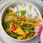 A bowl of green curry tofu served with white rice. A hand holding chopsticks is picking up a piece of tofu.