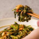 Chopsticks holding a portion of stir-fried rau muong with garlic and chili, above a plate of the same dish.