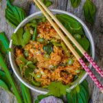 A bowl of Thai basil fried rice with vegetables and herbs, topped with sliced green onions and garnished with parsley, accompanied by two chopsticks.