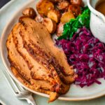 Plate with sliced vegan corned beef, roasted potatoes, sautéed greens, red cabbage, and a gravy boat.