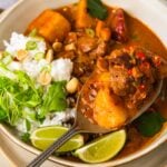 A bowl of vegan massaman curry with chunks of seitan, potatoes, and red chilies, served with rice, peanuts, cilantro, and lime wedges.