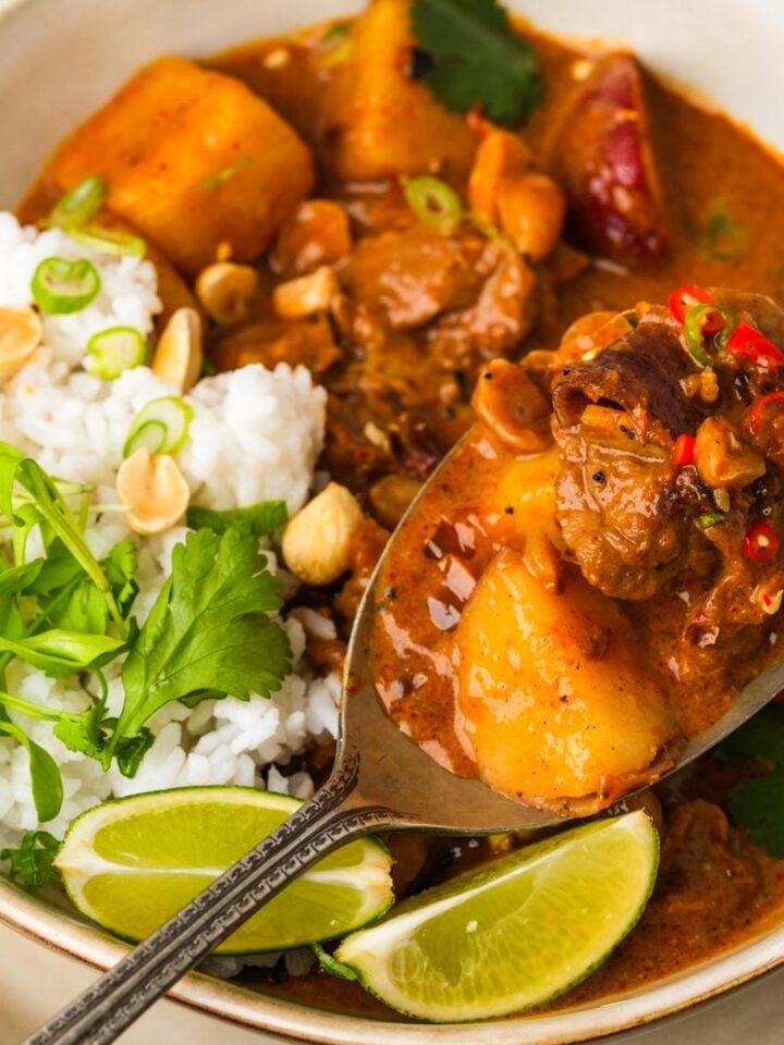 A bowl of vegan massaman curry with chunks of seitan, potatoes, and red chilies, served with rice, peanuts, cilantro, and lime wedges.