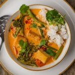 A bowl of vegan red curry with tofu, broccolini, and green beans, served with a portion of white rice and garnished with cilantro. A spoon rests on the side of the plate.