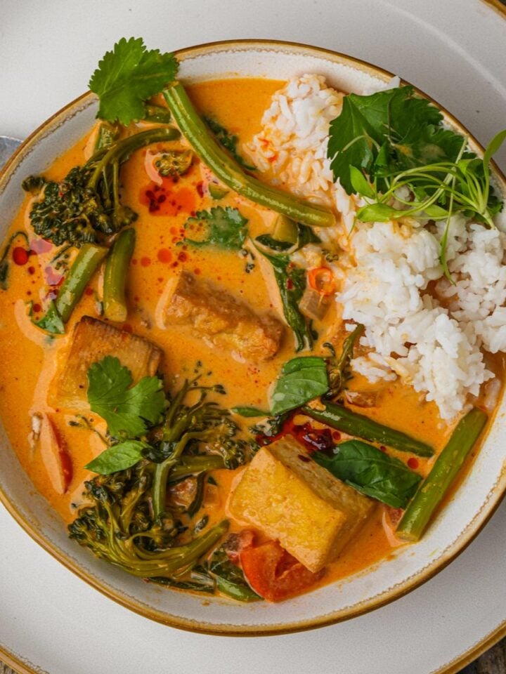 A bowl of vegan red curry with tofu, broccolini, and green beans, served with a portion of white rice and garnished with cilantro. A spoon rests on the side of the plate.