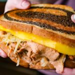 Two hands hold a vegan Reuben sandwich with marbled rye bread, vegan corned beef, sauerkraut, and melted cheese.