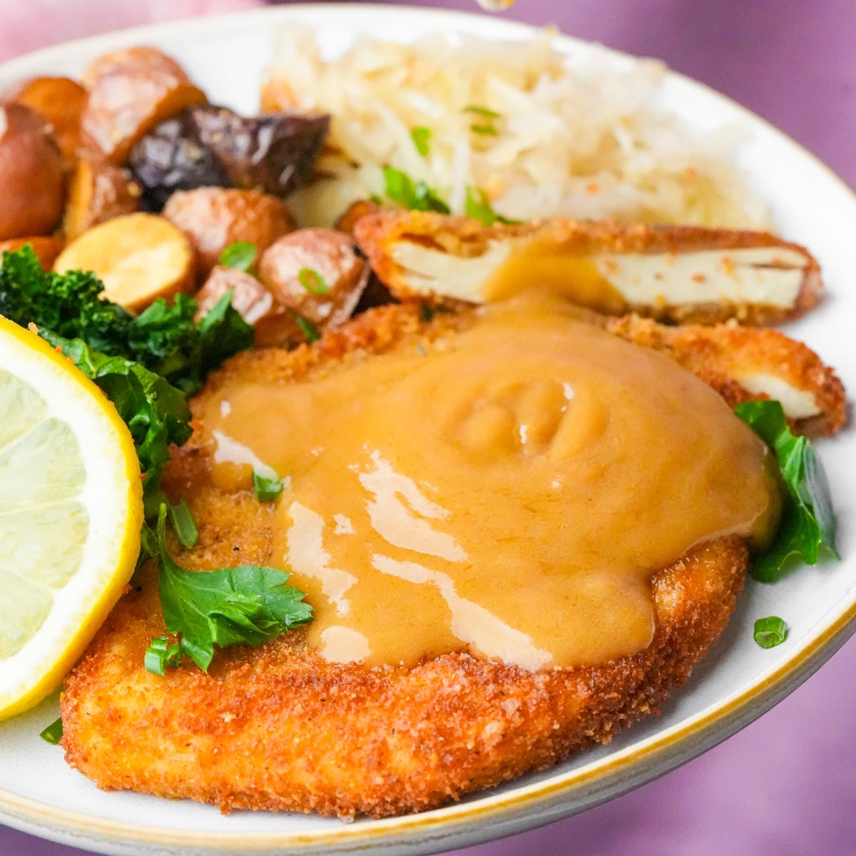 vegan_schnitzel-01 A plate with breaded vegan schnitzel topped with brown gravy, a lemon slice, roasted potatoes, sauerkraut, and greens on the side.