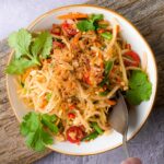 A bowl of Thai som tam with shredded green papaya, cherry tomatoes, and garnished with cilantro and fried shallots. A spoon is partially visible on the side.