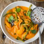A bowl of spicy vegan yellow curry with tofu, green beans, and cilantro, accompanied by a metal slotted spoon on a wooden surface.