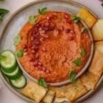 A bowl of muhammara topped with pomegranate seeds and parsley, served with cucumber slices and pita chips on a plate.