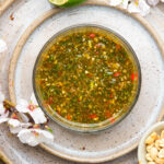A glass bowl of green chimichurri sauce with visible herbs and red chili flakes, surrounded by white flowers, a squeezed lime half, and a small bowl of peanuts on a speckled plate.