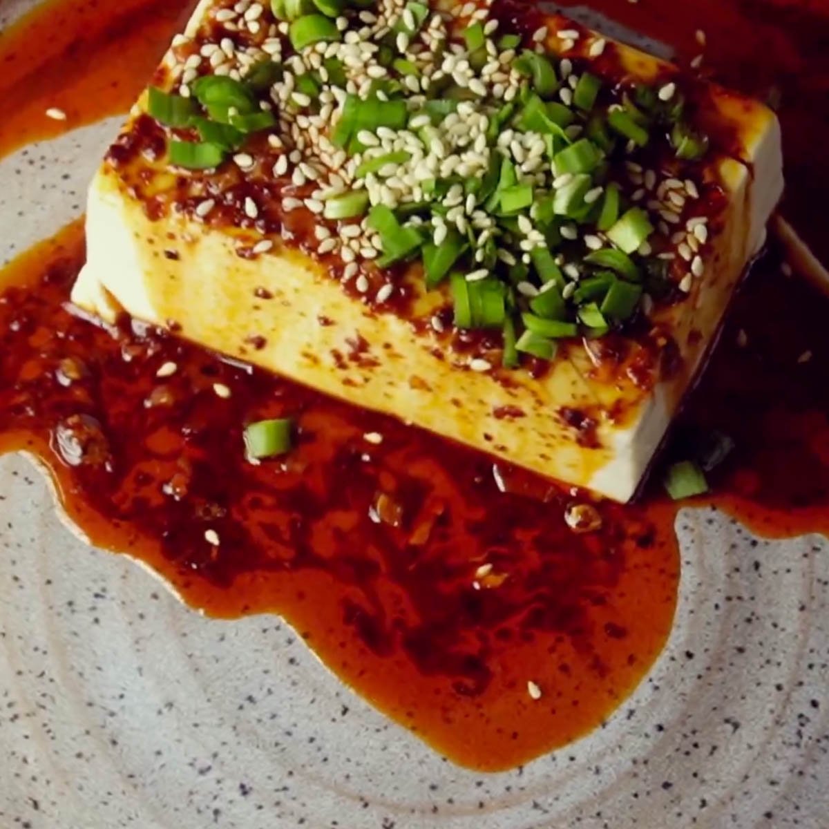 A block of tofu topped with green onions and sesame seeds, surrounded by a spicy red sauce on a gray speckled plate.