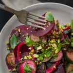 A fork holds a slice of beet above a salad with beets, pistachios, herbs, and greens on a plate.