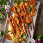 Roasted harissa carrots served on a bed of yogurt, garnished with chopped pistachios, parsley, and apricots, presented on a rectangular plate with flowers around it.