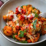 A bowl of harissa cauliflower florets topped with a creamy tahini sauce, garnished with fresh herbs.