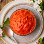 A glass bowl filled with red harissa sauce sits on a ceramic plate next to a spoon, with white flowers and green leaves as decoration.