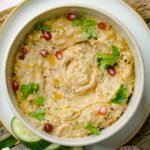 A bowl of Lebanese baba ganoush garnished with fresh parsley, pomegranate seeds, olive oil, and black pepper, served with cucumber slices on the side.