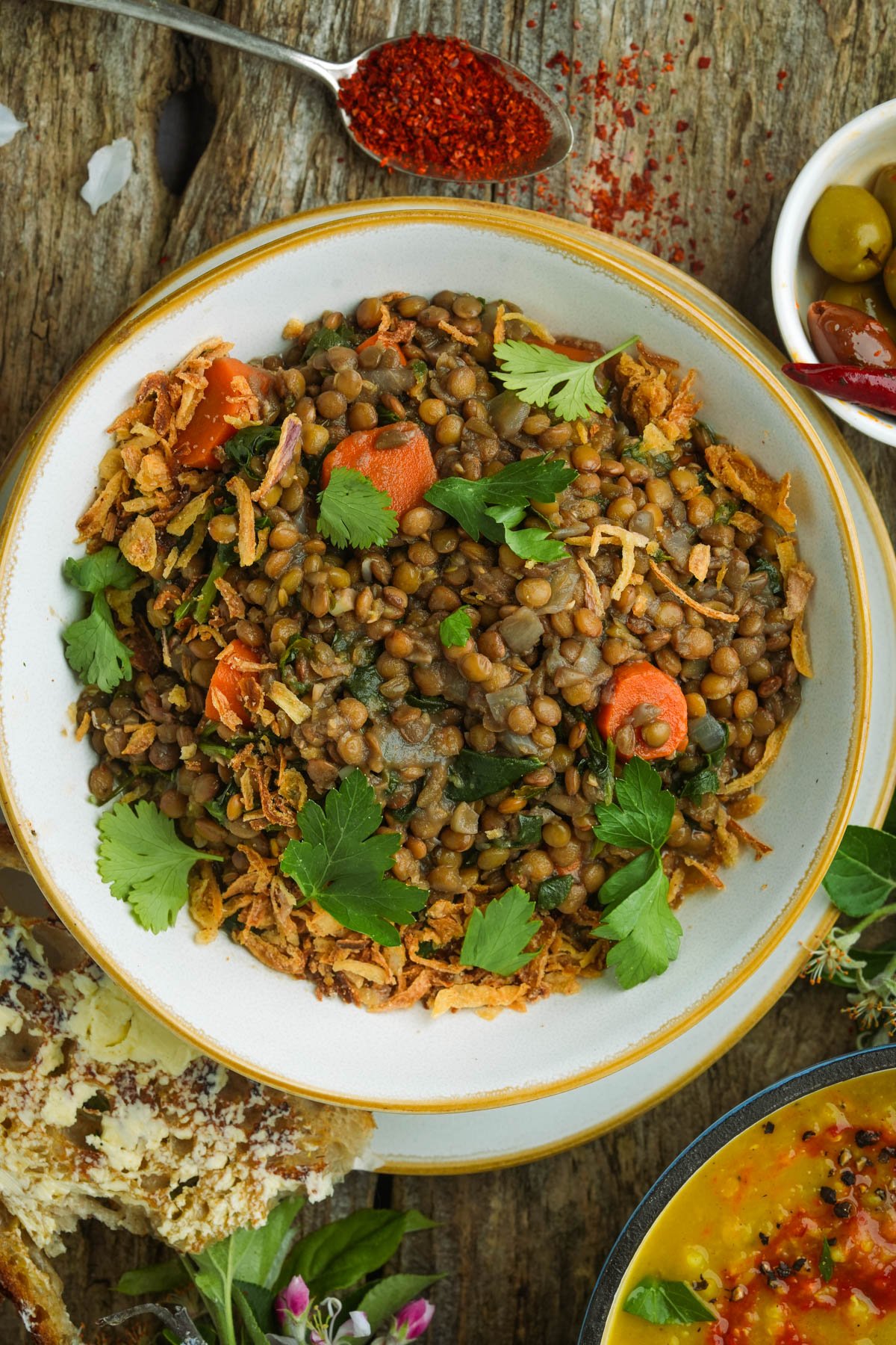 A bowl of Moroccan lentils garnished with fresh herbs and sliced carrots, placed on a rustic wooden surface with spices and olives nearby.