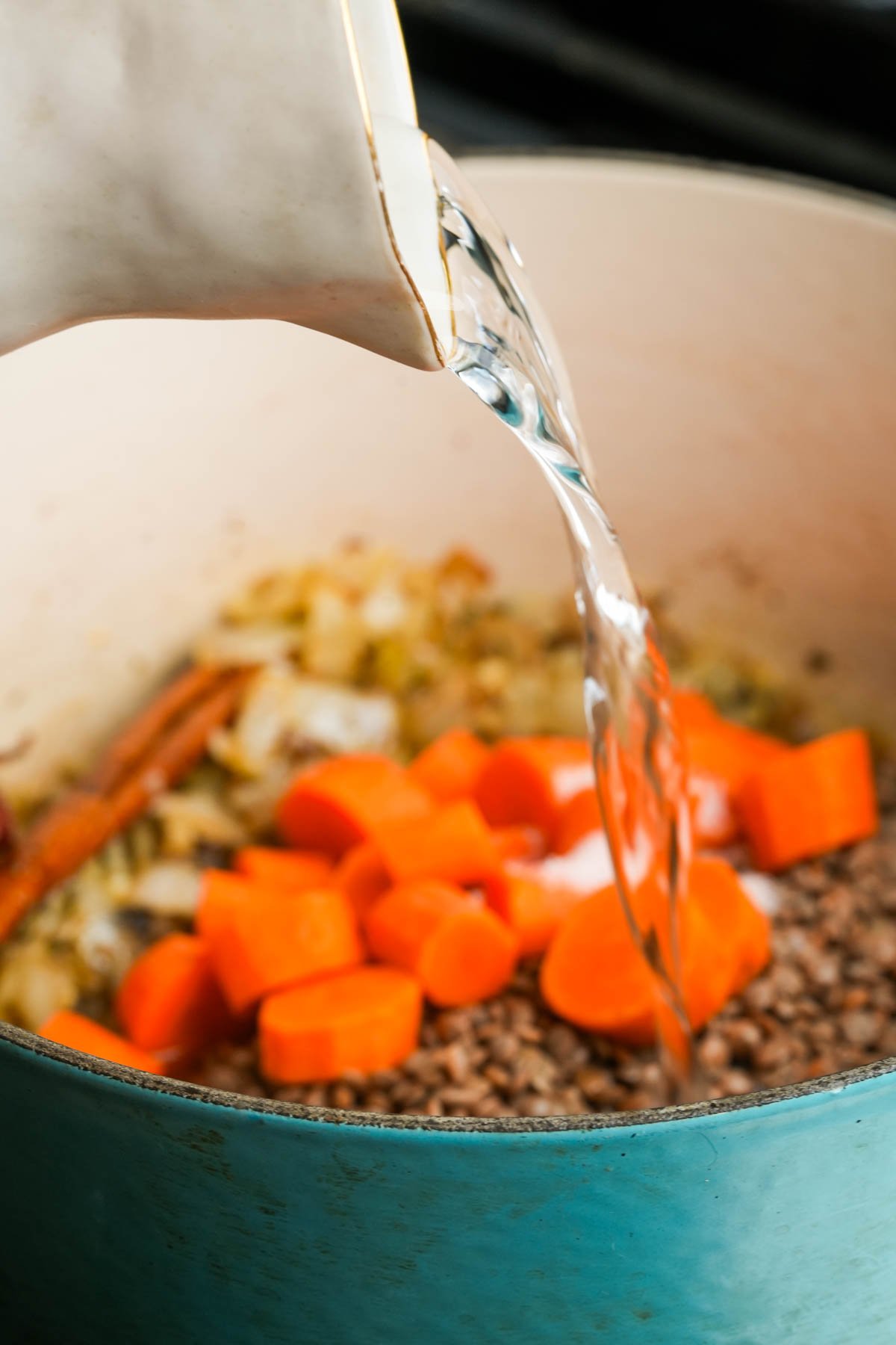 Water being poured from a pitcher into a pot containing chopped carrots, onions, and Moroccan lentils.