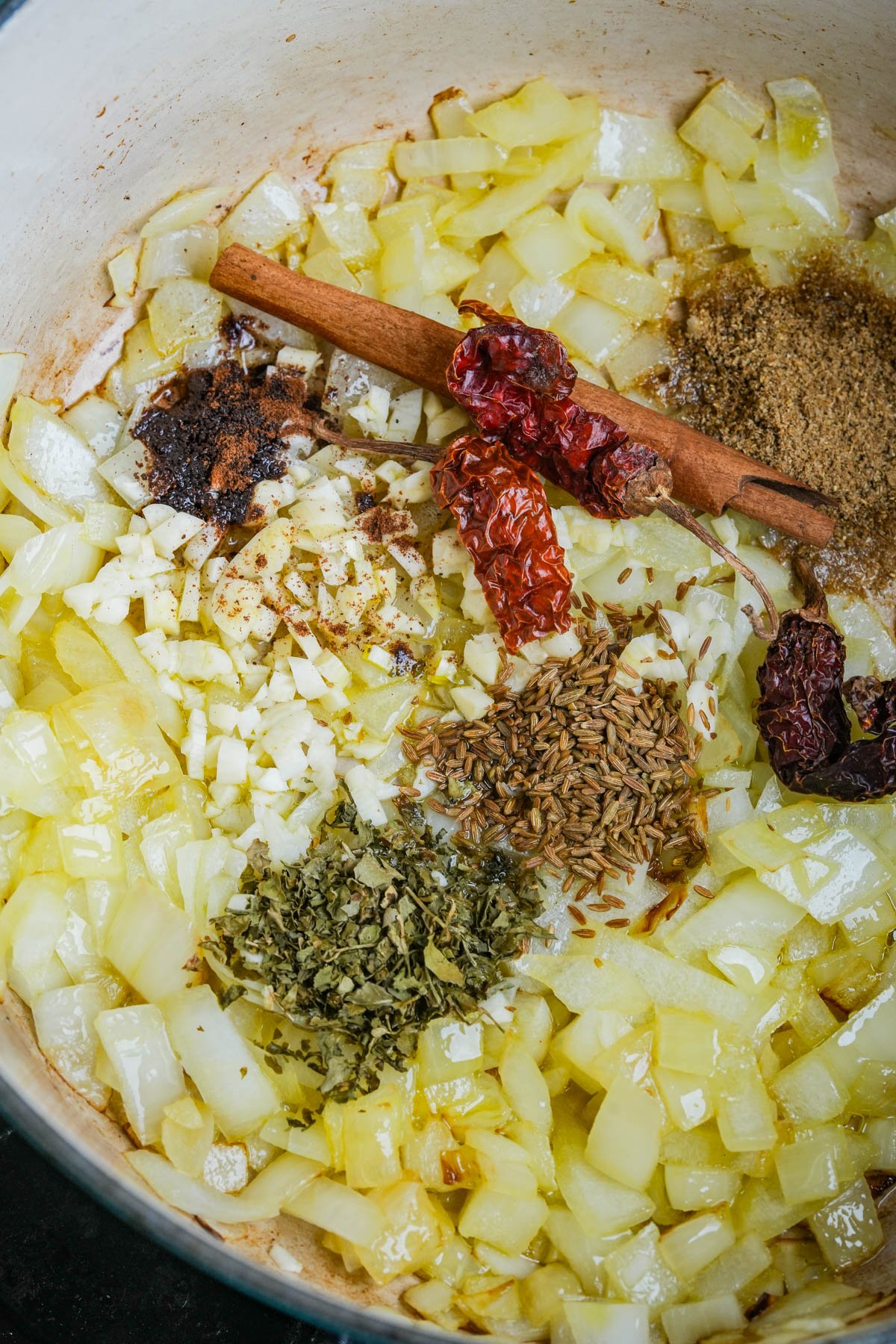 Chopped onions cooking in oil with garlic, dried red chilies, a cinnamon stick, spices, and herbs in a pot.