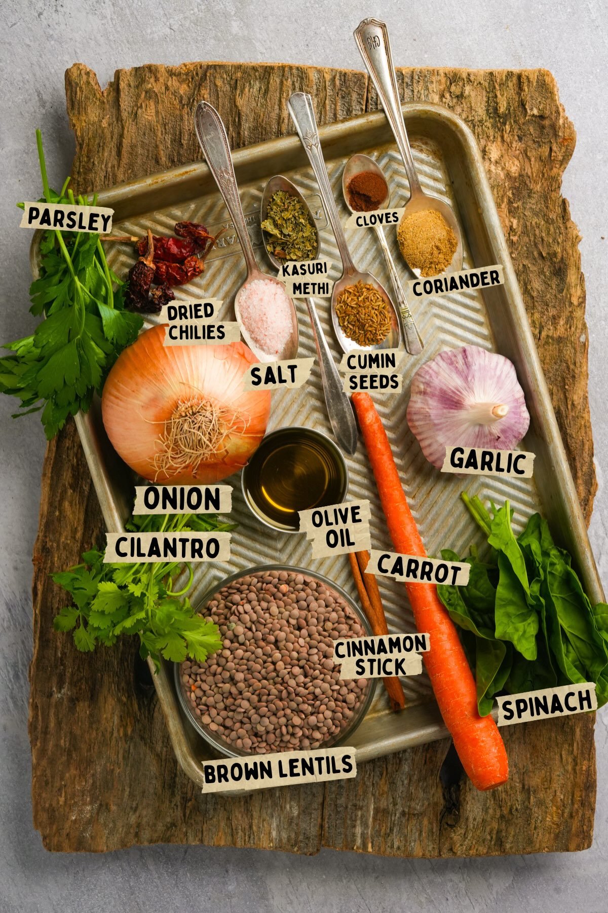 A tray on a wooden board holds labeled ingredients for a Moroccan lentils recipe: parsley, dried chilies, kasuri methi, cloves, coriander, onion, salt, cumin seeds, garlic, cilantro, olive oil, carrot, cinnamon stick, brown lentils, spinach.