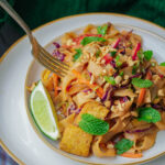 A close-up of a plate of vegetable pad Thai with tofu, peanuts, lime wedge, and fresh mint leaves, garnished with chopped vegetables and a fork on the side.