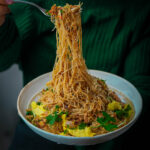 A person holds up a forkful of thin noodles from a bowl. The noodles are mixed with vegetables and garnished with cilantro. A green sweater is visible in the background.