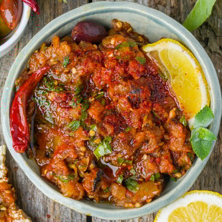 A bowl of zaalouk garnished with fresh herbs, a dried red chili, olives, and a lemon wedge, placed on a rustic wooden surface.