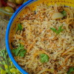A bowl of Egyptian rice topped with caramelized onions and garnished with parsley, with a side dish of olives visible in the background.