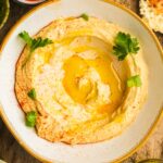 A bowl of Israeli hummus drizzled with olive oil, garnished with parsley and sprinkled with paprika on a rustic wooden surface.
