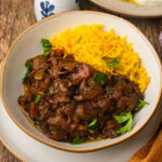 A plate with yellow rice and Khoresh bademjan garnished with parsley, served on a rustic wooden table.