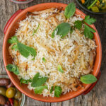 A bowl of cooked Lebanese rice garnished with fried onions, mint leaves, and parsley, placed on a wooden surface with side dishes nearby.