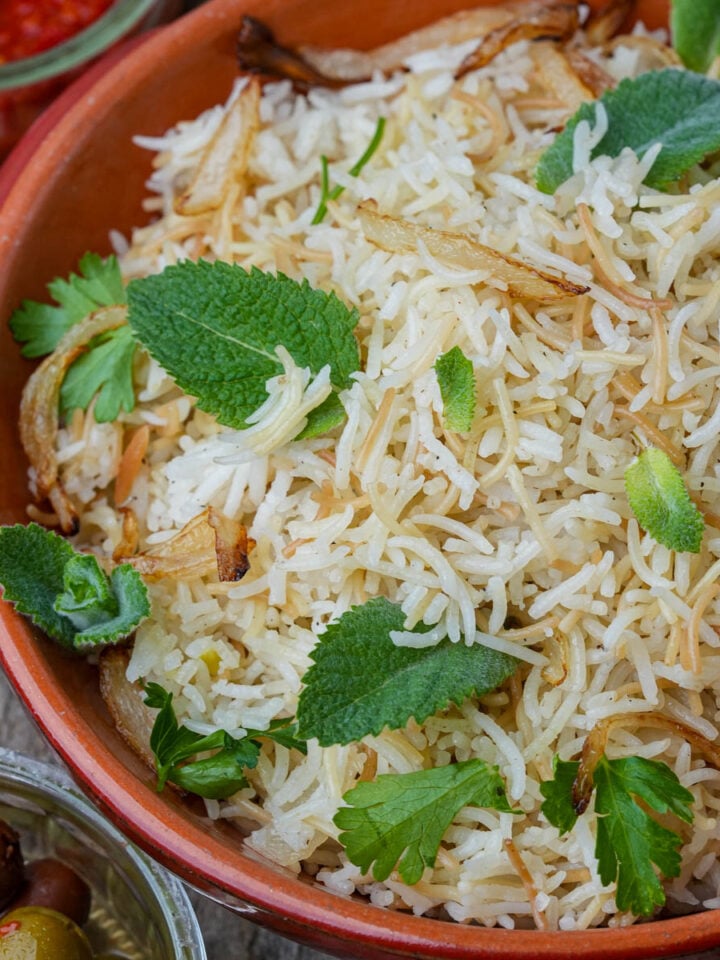 A bowl of Lebanese rice garnished with fried onions, fresh mint, and parsley, with a side of olives and a red sauce visible nearby.